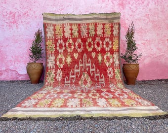 Puede incluir: Una alfombra marroquí roja, verde y blanca con un patrón geométrico. La alfombra está flecos y tiene un aspecto desgastado y vintage.