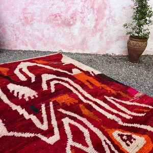 May include: A vibrant, hand-woven rug with a bold geometric pattern in shades of red, orange, and white. The rug's design features abstract shapes and lines, creating a striking visual effect. The rug is placed on a gravel surface, with potted plants in the background.