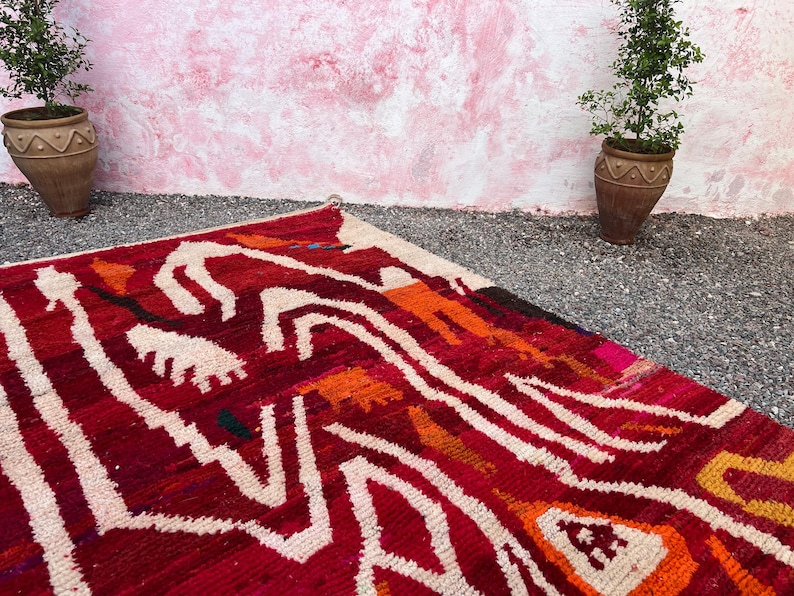 May include: A vibrant, hand-woven rug with a bold geometric pattern in shades of red, orange, and white. The rug's design features abstract shapes and lines, creating a striking visual effect. The rug is placed on a gravel surface, with potted plants in the background.