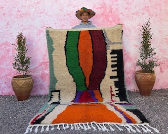 May include: A colorful, hand-woven rug with a geometric design. The rug features a cream-colored background with vertical stripes of green, orange, and burgundy. Black and white fringe lines the bottom edge. The rug is displayed outdoors.