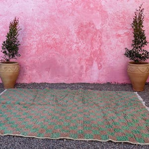 May include: A green and brown woven rug with a checkered pattern. The rug is laid out on a gravel surface in front of a pink wall with two potted plants.
