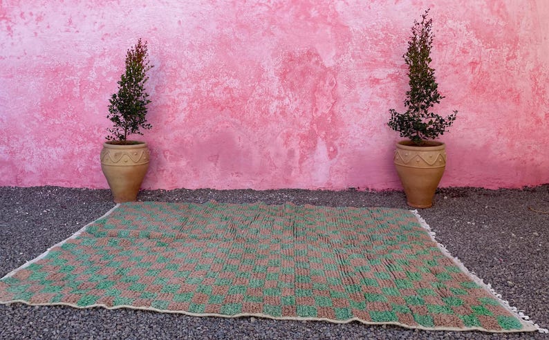 May include: A green and brown woven rug with a checkered pattern. The rug is laid out on a gravel surface in front of a pink wall with two potted plants.