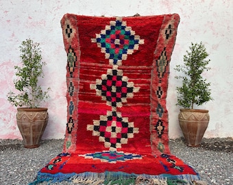 May include: A vibrant red Moroccan rug with geometric diamond patterns in black, white, blue, and green. The rug has a fringed edge and is displayed against a white wall, flanked by potted plants.