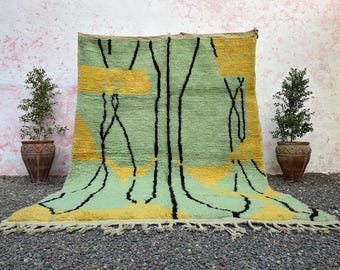 May include: A large, rectangular rug with a light green background and yellow geometric shapes. Black lines create a linear design. The rug has a fringe along the bottom edge. Two potted plants flank the rug.