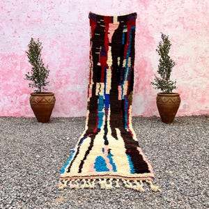 May include: A long, colorful rug with a geometric pattern in shades of blue, red, brown, and white. The rug has a fringe on both ends and is laid out on a gravel surface in front of a pink wall.