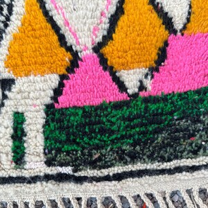 May include: Close-up of a colorful, textured rug with a geometric pattern. The rug features vibrant hues of pink, yellow, green, and black against a cream background. The design includes diamond and triangular shapes. The rug has a fringe along the bottom edge.