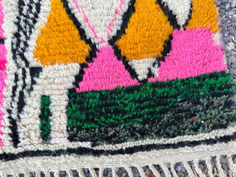 May include: Close-up of a colorful, textured rug with a geometric pattern. The rug features vibrant hues of pink, yellow, green, and black against a cream background. The design includes diamond and triangular shapes. The rug has a fringe along the bottom edge.
