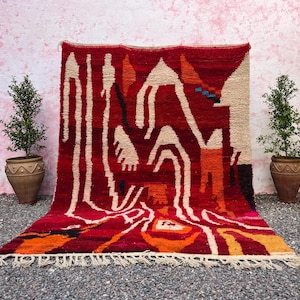 May include: A vibrant red rug with an abstract design featuring cream, orange, and black shapes. The rug has a fringed edge and is displayed against a pink wall, flanked by potted plants.