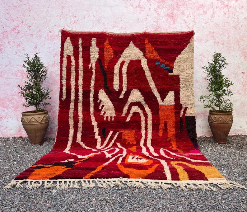 May include: A vibrant red rug with an abstract design featuring cream, orange, and black shapes. The rug has a fringed edge and is displayed against a pink wall, flanked by potted plants.