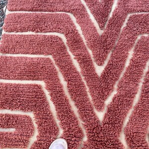 May include: A close-up of a pink and white patterned rug with a geometric design. The rug is made of a soft, fluffy material.