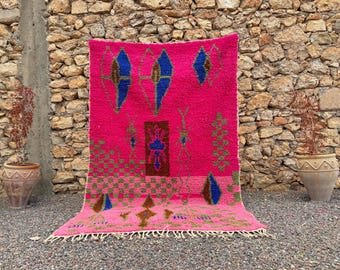 May include: A rectangular area rug featuring a vibrant hot pink background with a variety of geometric patterns in blue, brown, and green. The design includes diamond shapes, checkered sections, and vertical motifs, finished with a white fringe edge.