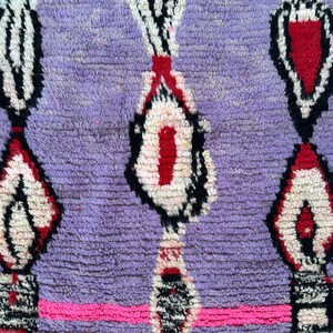 May include: Close-up of a hand-woven rug with a textured, purple background. The rug features a repeating pattern of white, red, and black geometric shapes. A bright pink horizontal stripe runs across the bottom.