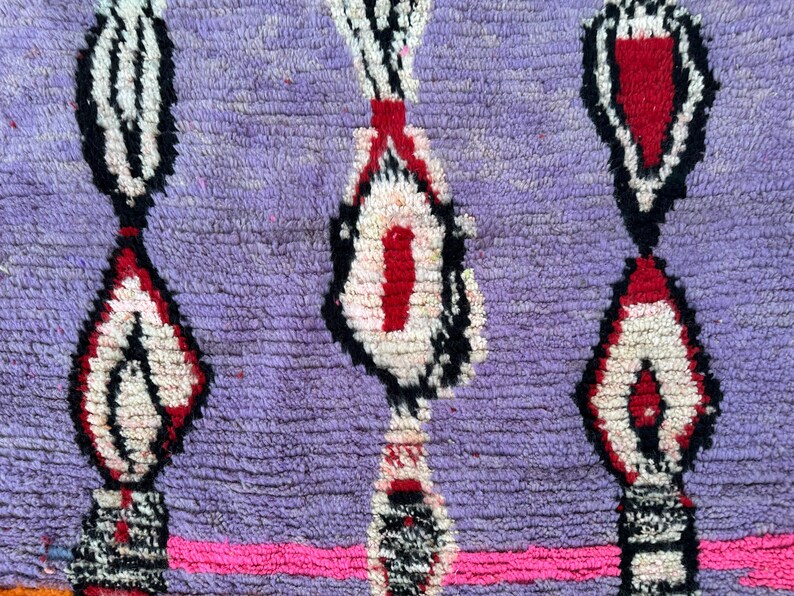 May include: Close-up of a hand-woven rug with a textured, purple background. The rug features a repeating pattern of white, red, and black geometric shapes. A bright pink horizontal stripe runs across the bottom.
