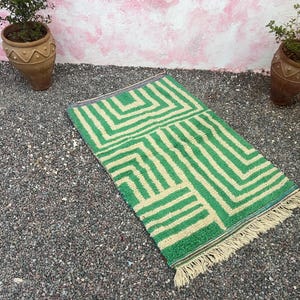 Pode incluir: Um tapete retangular verde e creme com um padr&atilde;o geom&eacute;trico. O tapete tem uma franja em uma extremidade e &eacute; colocado sobre uma cama de pequenas pedras cinzentas. Dois vasos de terracota com plantas est&atilde;o ao fundo.