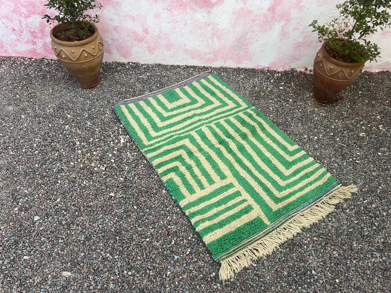 Pode incluir: Um tapete retangular verde e creme com um padr&atilde;o geom&eacute;trico. O tapete tem uma franja em uma extremidade e &eacute; colocado sobre uma cama de pequenas pedras cinzentas. Dois vasos de terracota com plantas est&atilde;o ao fundo.