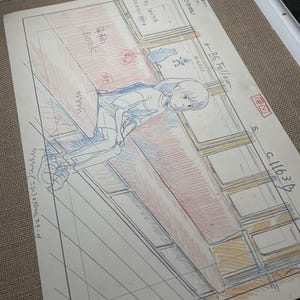 May include: A Studio Ghibli animation cel featuring a young girl in a blue and white outfit, seated near a window. The cel includes handwritten Japanese text and detailed pencil sketches, with red and blue color accents.