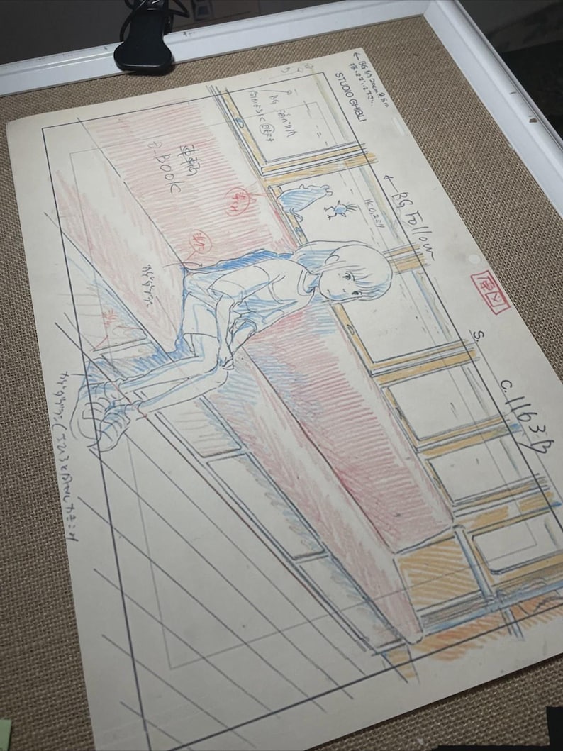 May include: A Studio Ghibli animation cel featuring a young girl in a blue and white outfit, seated near a window. The cel includes handwritten Japanese text and detailed pencil sketches, with red and blue color accents.