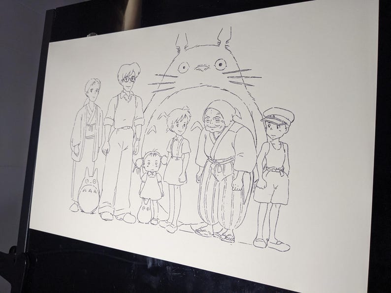 May include: A black and white line drawing of characters from the animated film 'My Neighbor Totoro'. The characters are standing in front of a large, furry creature with big eyes. The creature is Totoro, a friendly forest spirit.