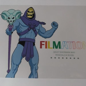 May include: Illustration of Skeletor, a blue-skinned character with a skull face, wearing a purple hood and belt. He holds a staff with a skull and ram's horns. The word "FILMATION" is in colorful letters.