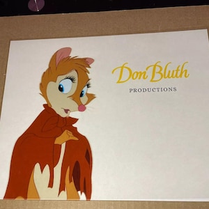 May include: A cel animation of a cartoon character with brown fur, wearing a red cloak. The character has large eyes and a pink nose. The image is signed "Don Bluth Productions".