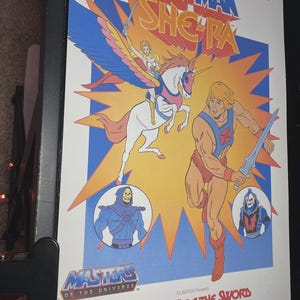 Puede incluir: Un p&oacute;ster enmarcado de la pel&iacute;cula animada "He-Man y She-Ra: El Secreto de la Espada". El p&oacute;ster presenta a He-Man, She-Ra y otros personajes en un dise&ntilde;o colorido y lleno de acci&oacute;n. El t&iacute;tulo se muestra de forma destacada.