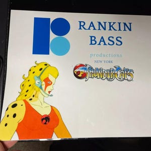 Puede incluir: Una tarjeta blanca con un logotipo azul y azul claro para las producciones de Rankin Bass, Nueva York. La tarjeta presenta una imagen de dibujos animados de Cheetara de ThunderCats.