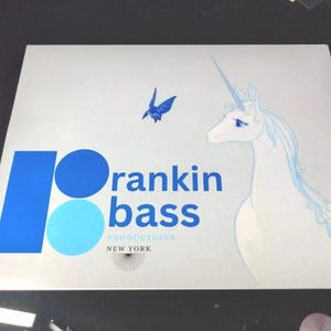 May include: A white card with blue and light blue circles and the text "Rankin Bass Productions New York". A white unicorn with a blue horn and a blue butterfly are also on the card.