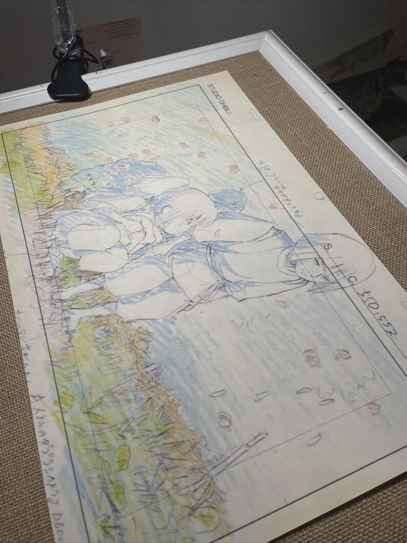 May include: A framed animation cel featuring a hand-drawn scene with two figures lying on the ground. The artwork is in blue and white pencil, with hints of color. The text "STUDIO GHIBLI" is visible in the upper right corner.