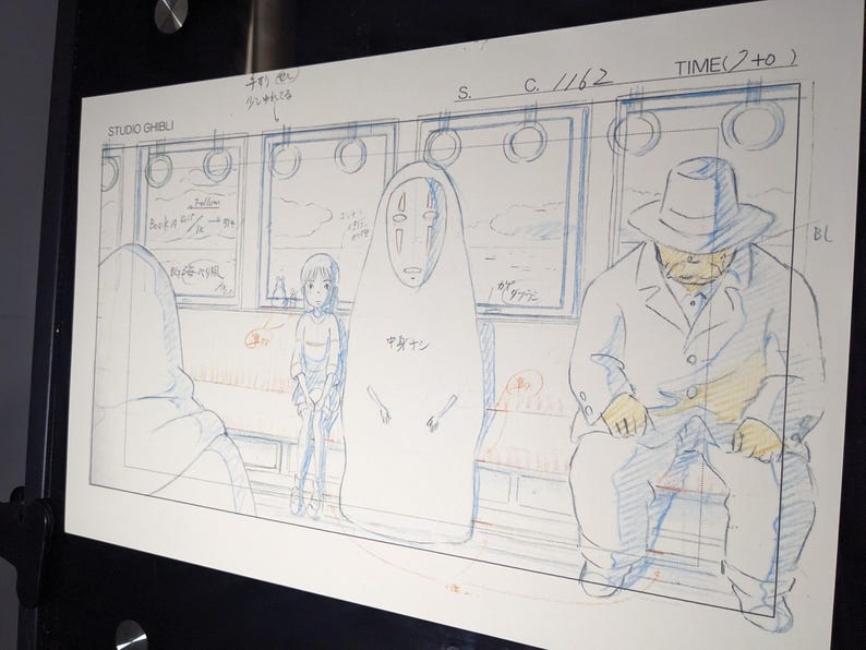 May include: A black and white hand-drawn illustration of three characters on a train. The characters are a young girl with short hair, a faceless spirit with a large, white, mask-like head, and a man wearing a hat and a coat. The illustration is on a white background with blue and red lines.