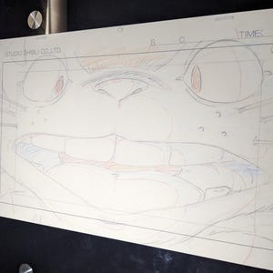 May include: A close-up of a pencil sketch of a cartoon character's face. The character has large eyes, a wide mouth, and a prominent nose. The sketch is on a white sheet of paper with a grid pattern.