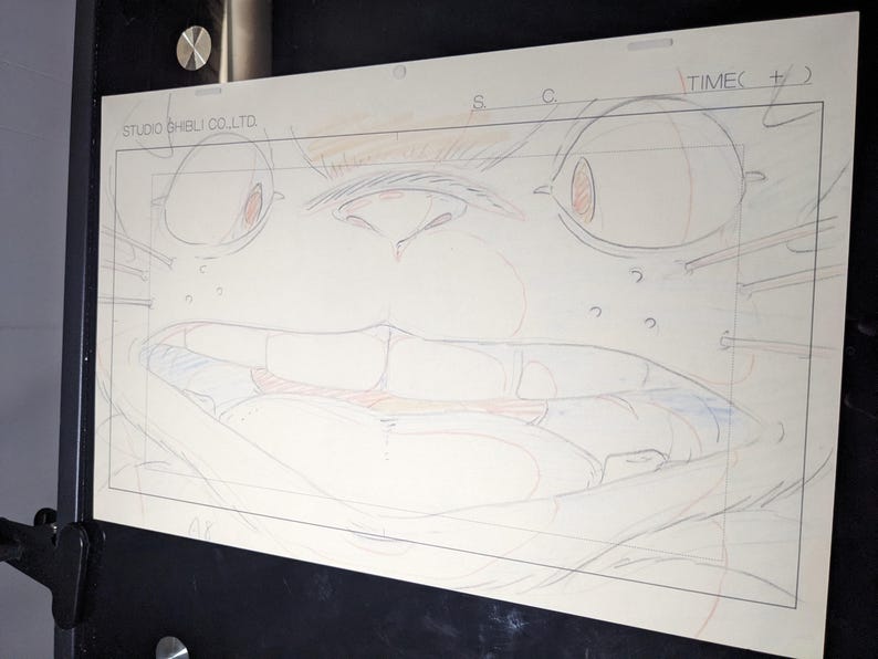 May include: A close-up of a pencil sketch of a cartoon character's face. The character has large eyes, a wide mouth, and a prominent nose. The sketch is on a white sheet of paper with a grid pattern.
