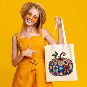 May include: A cream tote bag with a colourful, crocheted pumpkin design. The pumpkin is made of various square patches in shades of blue, green, pink, and yellow. The bag has a brown stem and green leaves.