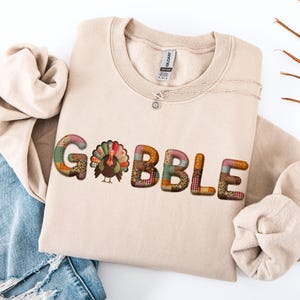 May include: A beige sweatshirt with the word "GOBBLE" in colourful patchwork letters. A cartoon turkey is featured in the centre. The sweatshirt is folded on top of a pair of blue jeans with distressed details.