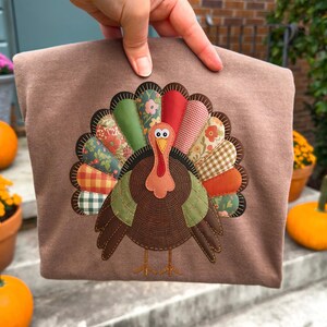 May include: A brown tote bag with a turkey applique. The turkey has a brown body, a red wattle, and a fan of patterned feathers in green, red, orange, and yellow. The bag is held up in front of a blurred background with pumpkins and flowers.