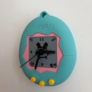 May include: A teal, egg-shaped wall clock with a pink square centre featuring the word Tamagotchi embossed at the top. The clock face has black hands, numbers 3, 6, 9, 12, and three yellow circular buttons at the bottom edge of the frame.