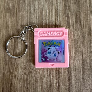 Puede incluir: Un llavero de cartucho de Gameboy rosa con el juego Pok&eacute;mon. El cartucho tiene la palabra "GAMEBOY" en la parte superior y una imagen de Jigglypuff. El llavero tiene un anillo y una cadena de metal plateado.