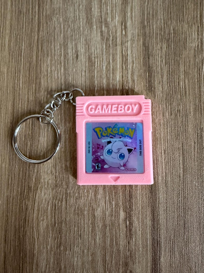 Puede incluir: Un llavero de cartucho de Gameboy rosa con el juego Pok&eacute;mon. El cartucho tiene la palabra "GAMEBOY" en la parte superior y una imagen de Jigglypuff. El llavero tiene un anillo y una cadena de metal plateado.