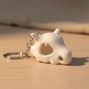May include: A white, 3D-printed keychain with a skull design, complete with a key ring and chain. The skull has large eye sockets and a rounded snout. The keychain is on a light brown wooden surface.
