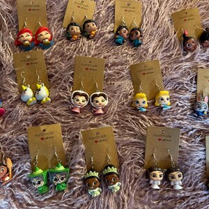 May include: A selection of handmade character earrings featuring Disney princesses, cartoon animals, and other figures. The earrings are attached to small, brown cardboard tags with the words "Handmade with love" printed on them. The earrings are displayed on a pink, fluffy surface.