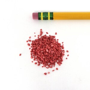 May include: A pile of small, red, crushed gravel. The gravel is scattered on a white surface. A yellow pencil with green stripes is lying above the gravel.