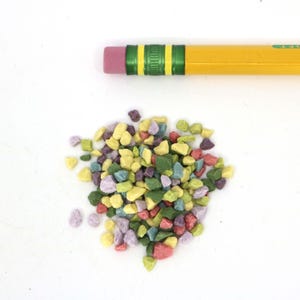 May include: A pile of colorful, small, decorative stones. The stones are in shades of yellow, green, purple, and red. The stones are scattered on a white surface.