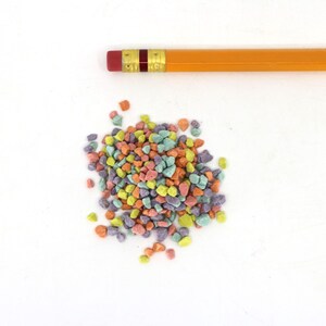 Könnte beinhalten: Ein Haufen bunter, unregelmäßig geformter Süßigkeiten in den Farbtönen Orange, Gelb, Grün, Rosa und Lila. Ein gelber Bleistift mit rotem Radiergummi liegt über den Süßigkeiten auf weißem Hintergrund.