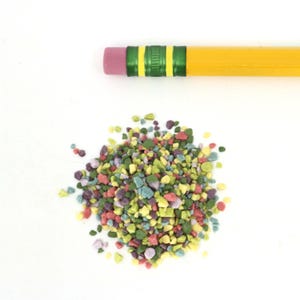 May include: A pile of colorful sprinkles in various shades of green, yellow, pink, and purple. The sprinkles are small and irregular in shape, resembling tiny pebbles.