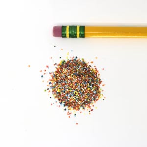 May include: A yellow pencil with a pink eraser and a small pile of colorful, granular material. The granular material is composed of various colors, including red, yellow, blue, and green. The pencil is positioned horizontally.