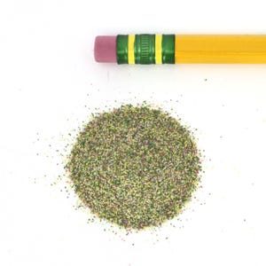 May include: A yellow pencil with a pink eraser and green and yellow accents rests above a pile of colorful, granular material. The material appears to be a mix of green, brown, and other colors, creating a textured appearance. The items are on a white background.