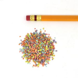Può includere: Un mucchio di piccole caramelle multicolori nelle tonalità di arancione, giallo, verde, viola e blu. Una matita gialla con una gomma rossa è posizionata sopra le caramelle. Lo sfondo è bianco.