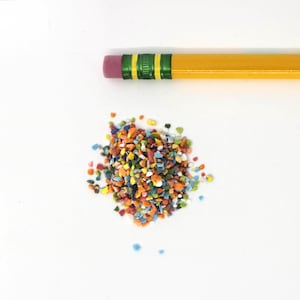 May include: A pile of colorful, small, irregular shaped pieces of confetti. The confetti is a mix of red, orange, yellow, green, blue, and white. A yellow pencil with a pink eraser is lying above the confetti.