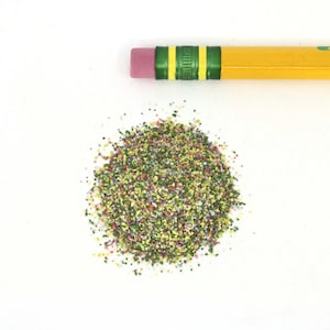 May include: A yellow pencil with a pink eraser and green stripes rests above a pile of colorful, granular material. The material is a mix of green, yellow, red, and blue particles, creating a textured appearance. The background is plain white.