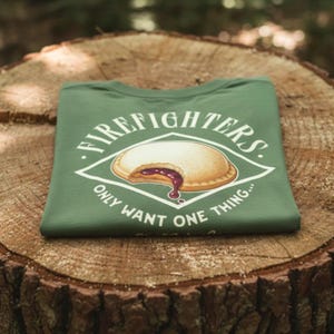 A folded green T-shirt with a vintage firefighter graphic featuring a bitten uncrustable snack and humorous text, displayed on a tree stump in a forest setting.
