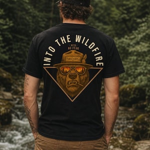 May include: A black t-shirt featuring a graphic on the back of a bear wearing a ranger hat and sunglasses with a forest fire reflection. The text reads INTO THE WILDFIRE WITH NO FEAR and Pine&EmberCo. The bear is framed by a triangle.